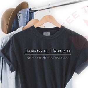 Pode incluir: Camiseta azul marinho escuro com as palavras "JACKSONVILLE UNIVERSITY" em letras brancas em bloco acima de "Business Administration" em fonte cursiva. A camiseta est&aacute; pendurada em um cabide de madeira com outras roupas.