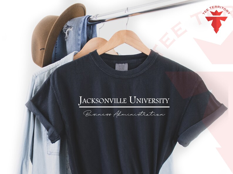Pode incluir: Camiseta azul marinho escuro com as palavras "JACKSONVILLE UNIVERSITY" em letras brancas em bloco acima de "Business Administration" em fonte cursiva. A camiseta est&aacute; pendurada em um cabide de madeira com outras roupas.