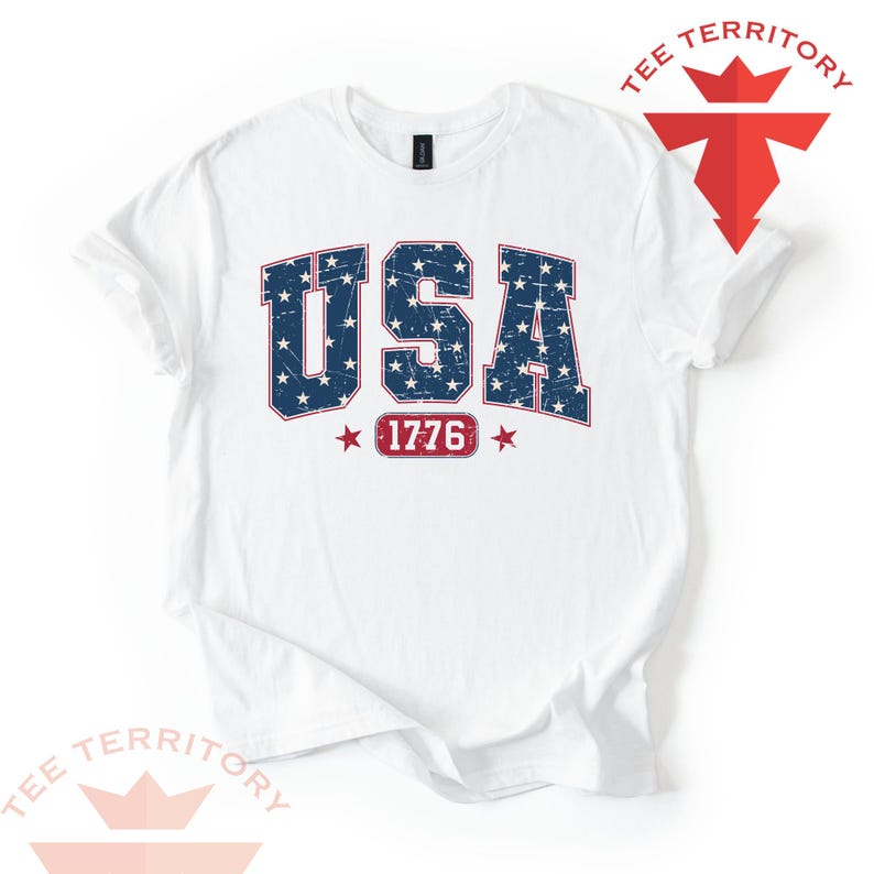 May include: White t-shirt with a distressed design featuring the word "USA" in blue with white stars, and the number "1776" in red. The shirt has a crew neck and short sleeves.