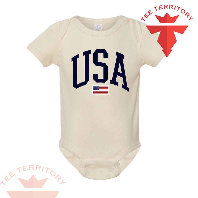 Puede incluir: Body de beb&eacute; color crema con la palabra "USA" en letras de bloque azul marino y una peque&ntilde;a bandera americana. El body tiene mangas cortas y cierre de broche.