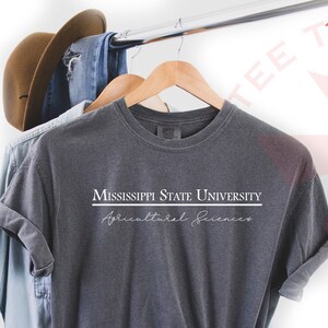 Pode incluir: Camiseta cinza escuro com o texto "MISSISSIPPI STATE UNIVERSITY Agricultural Sciences" em branco. A camisa est&aacute; pendurada em um cabide de madeira com um chap&eacute;u marrom e jeans azul ao fundo.
