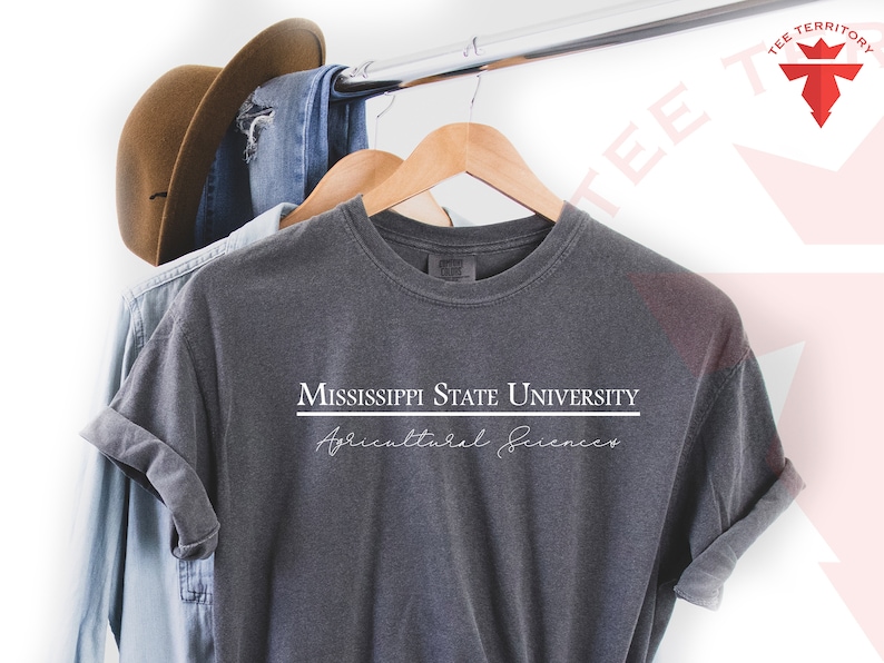 Pode incluir: Camiseta cinza escuro com o texto "MISSISSIPPI STATE UNIVERSITY Agricultural Sciences" em branco. A camisa est&aacute; pendurada em um cabide de madeira com um chap&eacute;u marrom e jeans azul ao fundo.