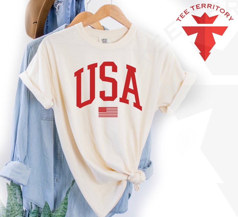 May include: A cream colored t-shirt with a red USA graphic and a small American flag. The shirt is on a hanger and is folded in half.