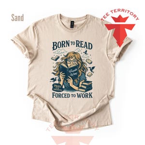 May include: Sand-colored t-shirt with a graphic that reads "Born to Read, Forced to Work." The illustration features a person reading a book with the title "Fourth Wing," surrounded by books and dragons. The shirt has short sleeves.