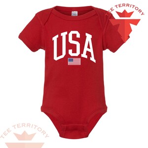 Puede incluir: Un body rojo para beb&eacute; con la palabra "USA" en letras blancas grandes. Debajo, una peque&ntilde;a bandera estadounidense. El body tiene mangas cortas y cierre de corchetes en la parte inferior. El texto "TEE TERRITORY" est&aacute; impreso arriba a la derecha e abajo a la izquierda.