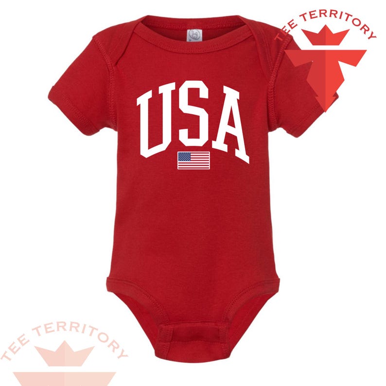 Puede incluir: Un body rojo para beb&eacute; con la palabra "USA" en letras blancas grandes. Debajo, una peque&ntilde;a bandera estadounidense. El body tiene mangas cortas y cierre de corchetes en la parte inferior. El texto "TEE TERRITORY" est&aacute; impreso arriba a la derecha e abajo a la izquierda.