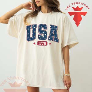 May include: Cream-colored t-shirt featuring the word "USA" in blue with white stars, and the number "1776" in a red banner. The shirt has a relaxed fit and short sleeves. The brand logo "TEE TERRITORY" is visible.