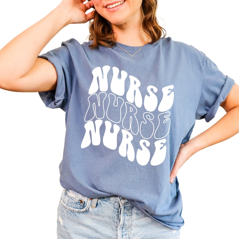 Puede incluir: Una camiseta azul con un gr&aacute;fico blanco de la palabra "NURSE" repetida tres veces en una fuente retro y ondulada.