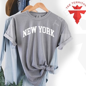 May include: Gray t-shirt with the text "NEW YORK" in white, displayed on a hanger. The shirt is made of a soft, comfortable fabric and has a relaxed fit.