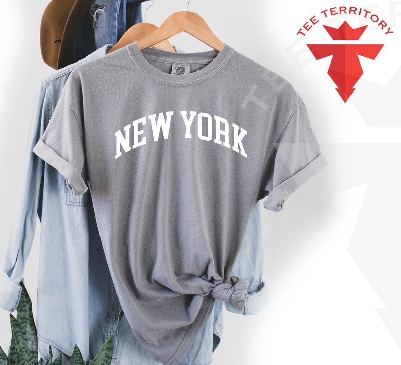 May include: Gray t-shirt with the text "NEW YORK" in white, displayed on a hanger. The shirt is made of a soft, comfortable fabric and has a relaxed fit.
