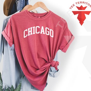 May include: A red t-shirt with the word "CHICAGO" printed in white on the front. The shirt is hanging on a wooden hanger.
