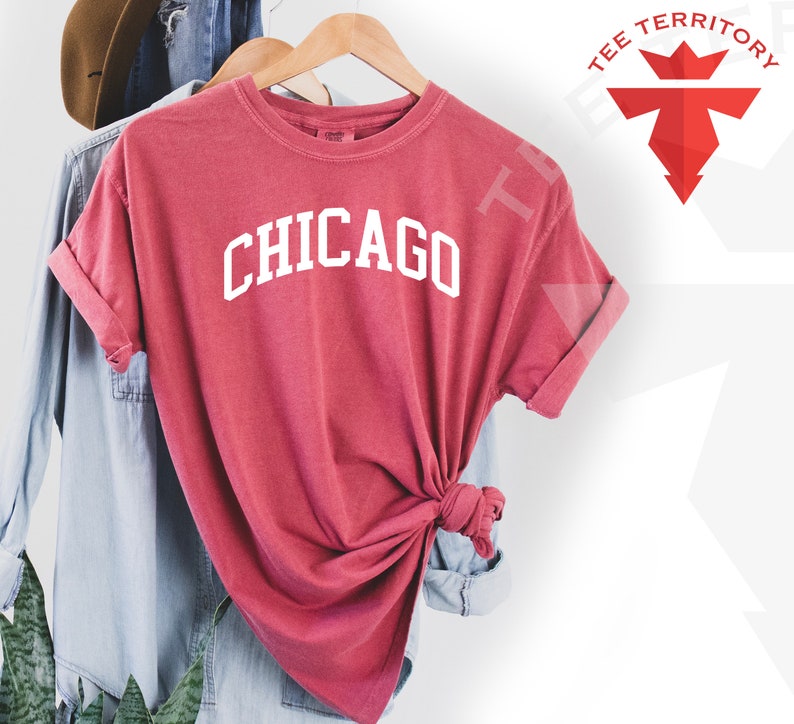 May include: A red t-shirt with the word "CHICAGO" printed in white on the front. The shirt is hanging on a wooden hanger.
