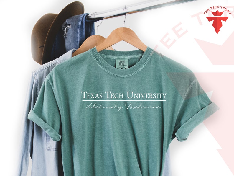 Pode incluir: Uma camiseta verde-azulado com o texto "Texas Tech University Veterinary Medicine" em branco. A camiseta est&aacute; pendurada em um cabide de madeira, com uma camisa azul clara e um chap&eacute;u marrom ao fundo.