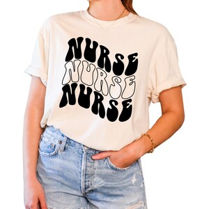 Puede incluir: Una camiseta blanca con un gr&aacute;fico negro de la palabra "NURSE" repetida tres veces en una fuente retro y ondulada.