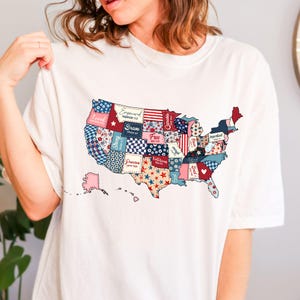 Pu&ograve; includere: Maglietta bianca con un design a patchwork della mappa degli Stati Uniti. Ogni stato &egrave; un motivo diverso con parole come "Strong", "Brave", "Free" e "Loved" su di essi.