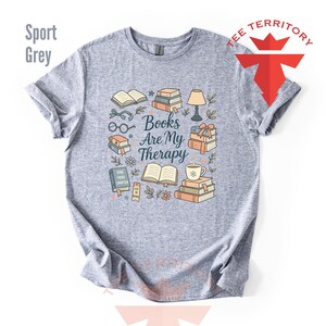 Może przedstawiać: Szara koszulka sportowa z kolorowym wzorem graficznym przedstawiającym książki, okulary, lampę i napis "Books Are My Therapy". Projekt zawiera filiżankę kawy, kwiaty i słowa "One More Chapter".