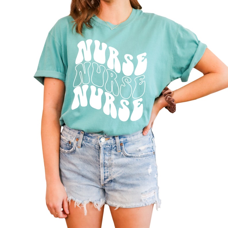 Puede incluir: Una camiseta azul turquesa con un gr&aacute;fico blanco que dice "NURSE" en una fuente retro y ondulada.
