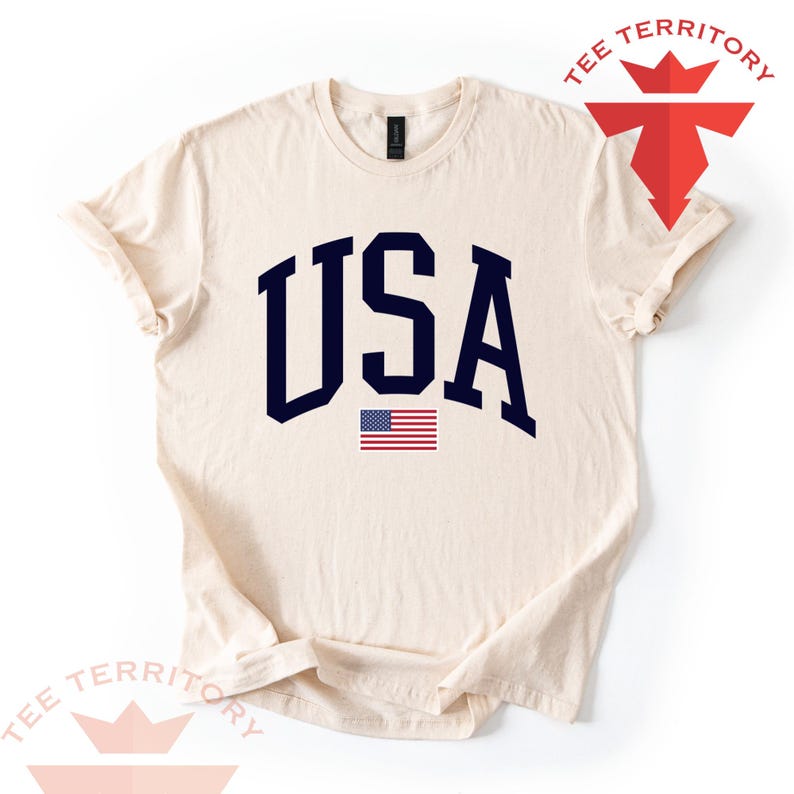 Puede incluir: Camiseta color crema con la palabra "USA" en letras de bloque azul marino y un peque&ntilde;o gr&aacute;fico de la bandera estadounidense. La camiseta tiene cuello redondo y mangas cortas. El logotipo de la marca "TEE TERRITORY" est&aacute; en la esquina superior derecha.