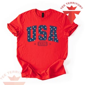 May include: A red t-shirt with the word "USA" in blue, filled with white stars. Below the word "USA" is the number "1776" in red, with a star on each side. The shirt has rolled-up sleeves.