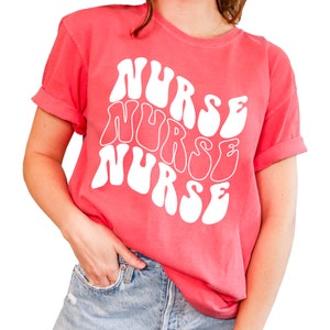 Puede incluir: Una camiseta rosa con la palabra "NURSE" repetida tres veces en una fuente retro blanca.