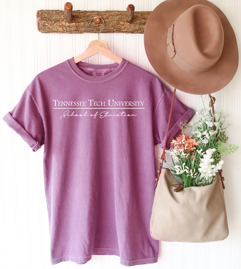 Pode incluir: Uma camiseta malva com mangas enroladas exibe o texto "Tennessee Tech University" acima de "School of Education" em branco. Um chap&eacute;u marrom e uma bolsa bege cheia de flores tamb&eacute;m s&atilde;o vis&iacute;veis. A camiseta est&aacute; pendurada em um cabide de madeira.