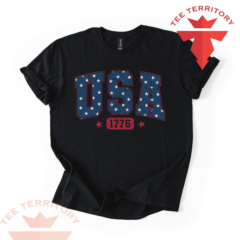 May include: Black t-shirt with the word "USA" in blue with white stars, and the number "1776" in red. The shirt has rolled-up sleeves and a crew neck. The shirt is from Tee Territory.
