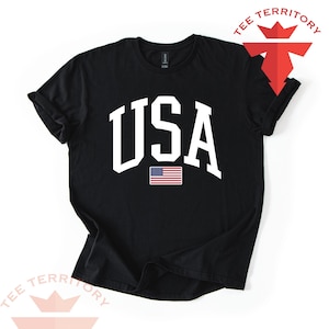 Puede incluir: Camiseta negra con la palabra "USA" en letras blancas grandes y un peque&ntilde;o gr&aacute;fico de la bandera estadounidense. La camiseta tiene las mangas enrolladas y las palabras "TEE TERRITORY" en rojo y blanco.
