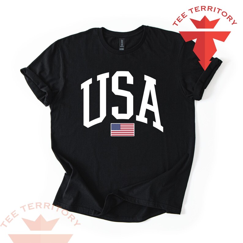 Puede incluir: Camiseta negra con la palabra "USA" en letras blancas grandes y un peque&ntilde;o gr&aacute;fico de la bandera estadounidense. La camiseta tiene las mangas enrolladas y las palabras "TEE TERRITORY" en rojo y blanco.