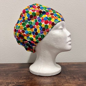 Puede incluir: Un gorro quirúrgico colorido con un patrón de lunares grandes y vibrantes en rojo, amarillo, verde, azul y morado sobre un fondo blanco. El gorro se muestra en una cabeza de maniquí blanca.