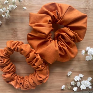 May include: Two orange scrunchies made of fabric, one large and one small, sit on a wooden surface. White flowers are scattered around the scrunchies. The scrunchies are a solid orange color and appear to be handmade.
