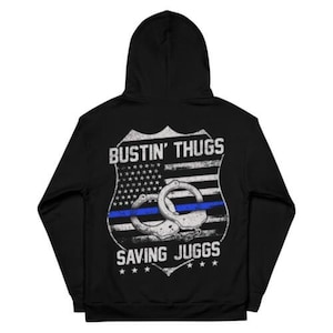 May include: Black hoodie with a white and blue graphic on the back. The graphic features a shield with an American flag, handcuffs, and the text "Bustin' Thugs Saving Juggs".