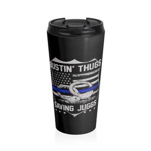 May include: Black travel mug with a white graphic of a police badge with an American flag, handcuffs, and the text "Bustin' Thugs Saving Juggs".
