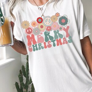 May include: White t-shirt with a retro-style "Merry Christmas" graphic featuring colorful flowers and peace signs.