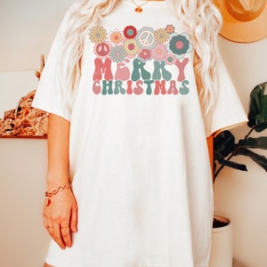 May include: White oversized t-shirt with a retro-style graphic design featuring the words "Merry Christmas" surrounded by colorful flowers and peace signs.