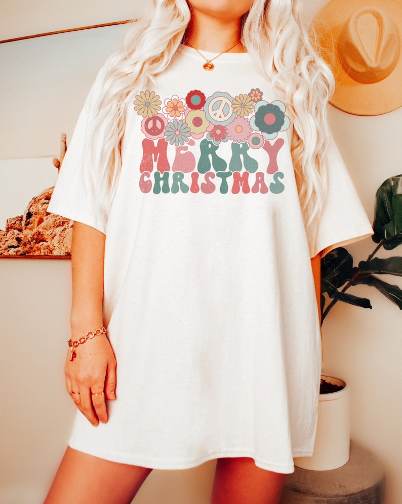 May include: White oversized t-shirt with a retro-style graphic design featuring the words "Merry Christmas" surrounded by colorful flowers and peace signs.