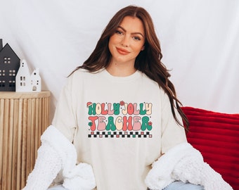 May include: A cream-colored sweatshirt with a festive design that reads "Holly Jolly Teacher" in a retro font with green, pink, and red colors. The design includes a Santa hat and flowers.