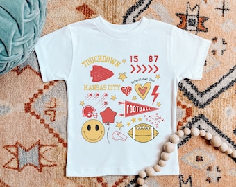 Puede incluir: Camiseta blanca con un diseño gráfico rojo y amarillo con las palabras "Touchdown", "Kansas City", "Football" y los números "15" y "87".