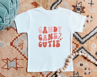 May include: White t-shirt with a pink and red graphic that says "Candy Cane Cutie" with a candy cane and star graphic.