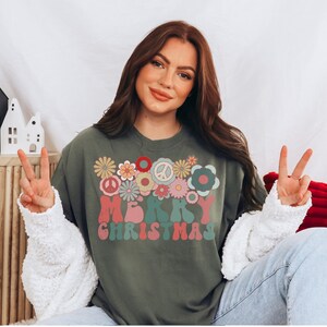 May include: A green long-sleeved sweatshirt with a retro-style "Merry Christmas" graphic featuring colorful flowers and peace signs.