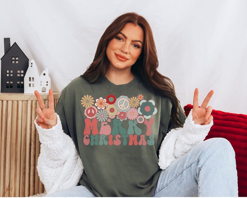May include: A green long-sleeved sweatshirt with a retro-style "Merry Christmas" graphic featuring colorful flowers and peace signs.