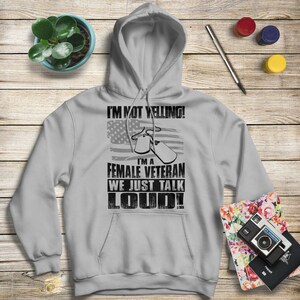 May include: A gray hooded sweatshirt with the text "I'm not yelling! I'm a female veteran we just talk loud!" printed on the front. The text is in black and is surrounded by a small American flag graphic and two dog tags.