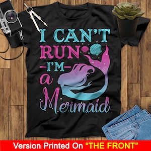May include: Black t-shirt with the text "I can't run - I'm a mermaid" in blue and pink lettering. A mermaid silhouette with a pink and blue gradient is featured on the shirt.