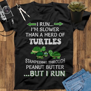 May include: Black t-shirt with a funny saying about running. The text reads "I run... I'm slower than a herd of turtles stampeding through peanut butter... but I run".