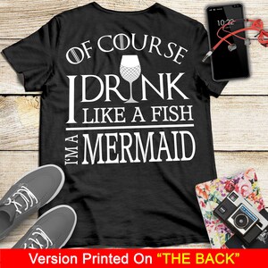 May include: Black t-shirt with white text that reads "Of course I drink like a fish I'm a mermaid". The text is surrounded by a white outline. A wine glass with a mermaid tail design is in the middle of the text.