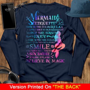 May include: A navy blue long-sleeved t-shirt with a mermaid graphic and the text "Mermaid Etiquette: Swim in the ocean relax, a little sand is okay, watch the sunset, enjoy, laugh with the gulls, gather seashells & wear pearls, smile, make friends with a starfish, sunbathe, splish splash, believe in magic".