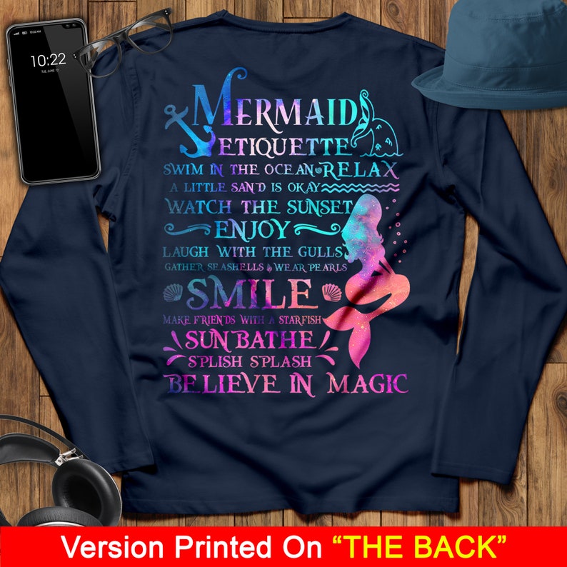 May include: A navy blue long-sleeved t-shirt with a mermaid graphic and the text "Mermaid Etiquette: Swim in the ocean relax, a little sand is okay, watch the sunset, enjoy, laugh with the gulls, gather seashells & wear pearls, smile, make friends with a starfish, sunbathe, splish splash, believe in magic".