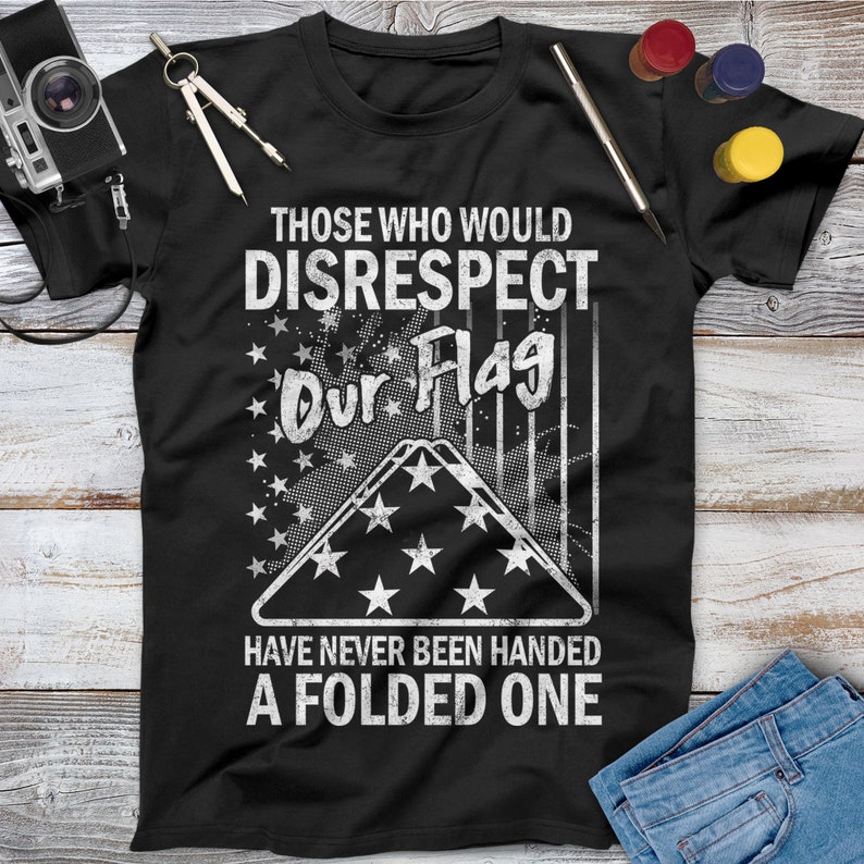 May include: Black t-shirt with a white graphic design. The graphic features a folded American flag with stars and stripes and the text "Those who would disrespect our flag have never been handed a folded one."