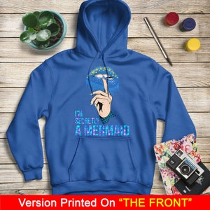 Peut inclure: Sweat-shirt &agrave; capuche bleu avec un graphique de l&egrave;vres bleues et violettes avec un doigt dessus, et le texte "I'm Secretly A Mermaid".