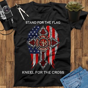 May include: Black t-shirt with a red, white, and blue American flag design. The flag is in the shape of a cross with a firefighter's emblem in the center. The text "Stand for the Flag" is at the top and "Kneel for the Cross" is at the bottom.
