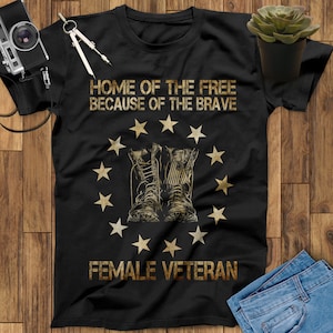 Puede incluir: Camiseta negra con un estampado dorado de un par de botas militares y estrellas. El texto "Home of the Free Because of the Brave" y "Female Veteran" est&aacute; impreso en la camiseta.